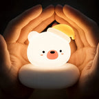 Veilleuse LED ours Bouba blanche avec bonnet jaune, allumée et diffusant une lumière chaude, entourée de deux mains.