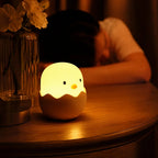 Veilleuse LED Coco en forme de poussin allumée sur une table de chevet en bois, à côté d'un vase de fleurs, dans une chambre tamisée le soir