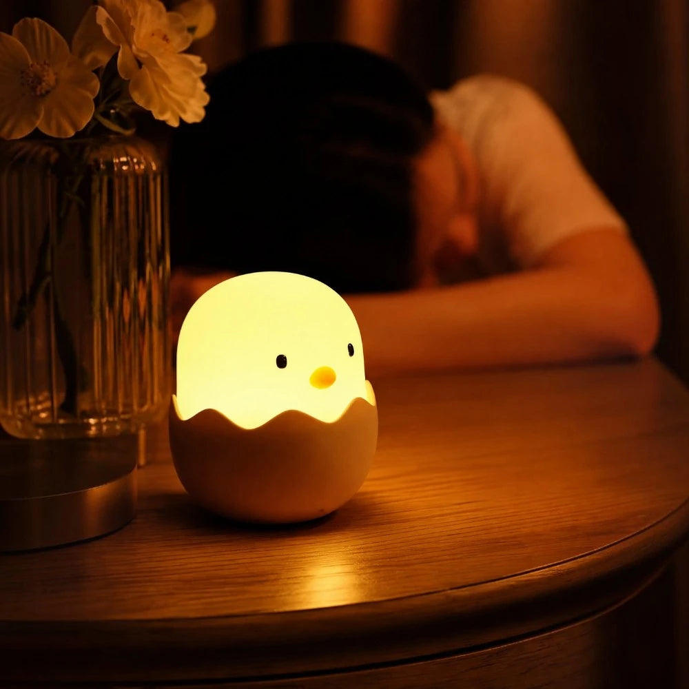 Veilleuse LED Coco en forme de poussin allumée sur une table de chevet en bois, à côté d'un vase de fleurs, dans une chambre tamisée le soir