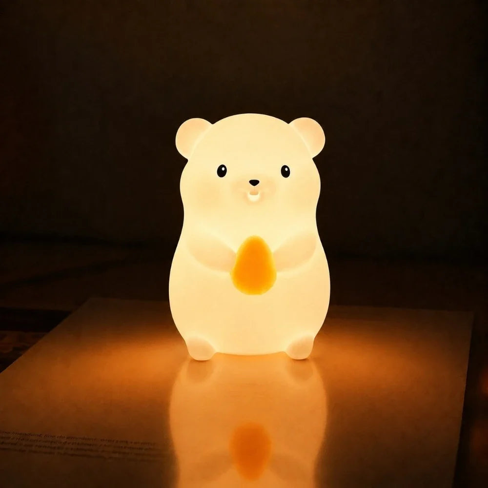 Veilleuse LED hamster Hamy allumée tenant une petite graine orange, diffusant une lumière chaude et apaisante