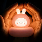 Veilleuse LED lapin Luco rose allumée, diffusant une lumière chaude et entourée de deux mains.
