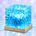 Veilleuse projecteur Aqua allumée en bleu, cube en acrylique texturé effet vagues sur socle effet bois, fond bleuté apaisant