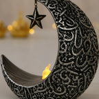 Vue 3/4 arrière zoomée veilleuse LED croissant de lune argentée, gravures calligraphie arabe en relief, étoile suspendue et flamme LED, fond bokeh doré